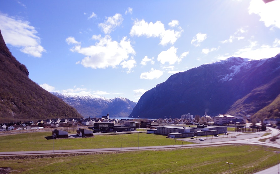 Utsikt mot Høyanger-sentrum med grønt gress, blå himmel og snødekte fjell. Alt med et lilla-skjær.