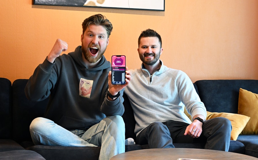 Supertrekning-vinner Joakim Norberg Klingenberg og broren Mikael sitter i en grå sofa i stuen til Joakim med oransje bakgrunn. Joakim holder opp mobilen med vinner-meldingen fra Norsk Tipping og jubler inn i kameraet.