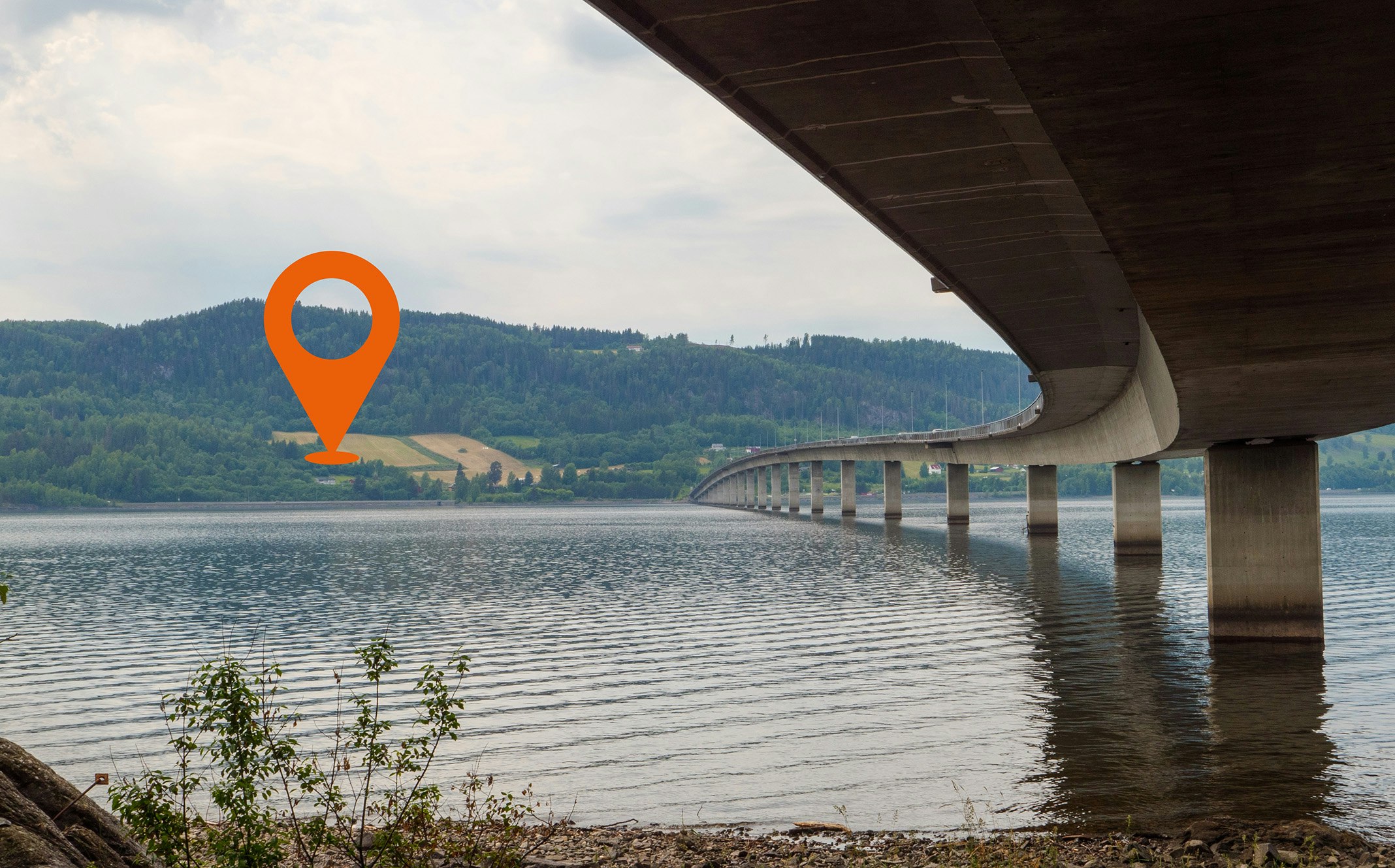 Bilde av Mjøsbrua underifra, med mjøsa i forgrunn og landskap i bakgrunnen.