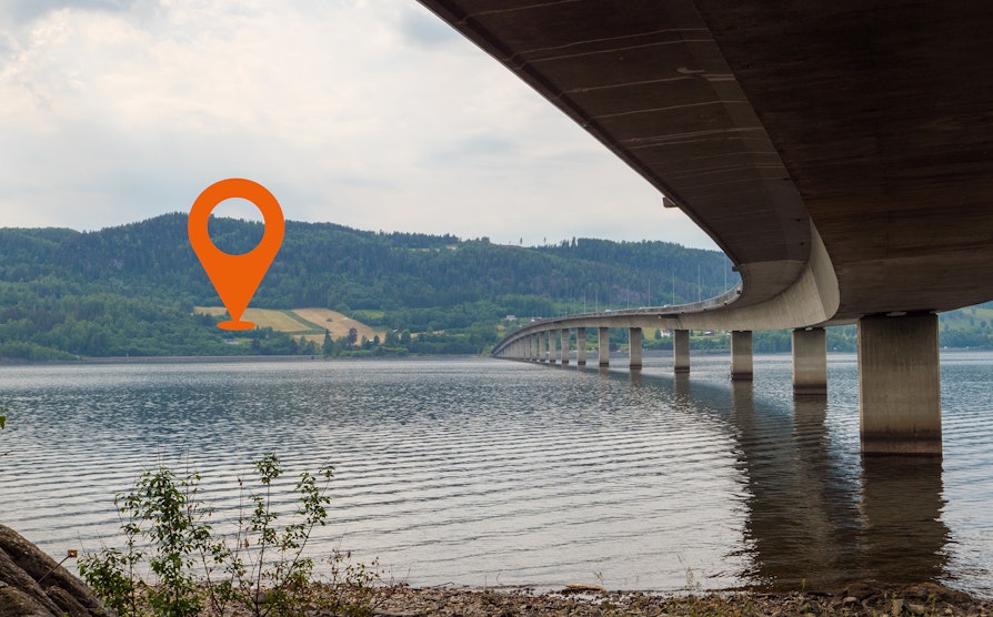Bilde av Mjøsbrua underifra, med mjøsa i forgrunn og landskap i bakgrunnen.
