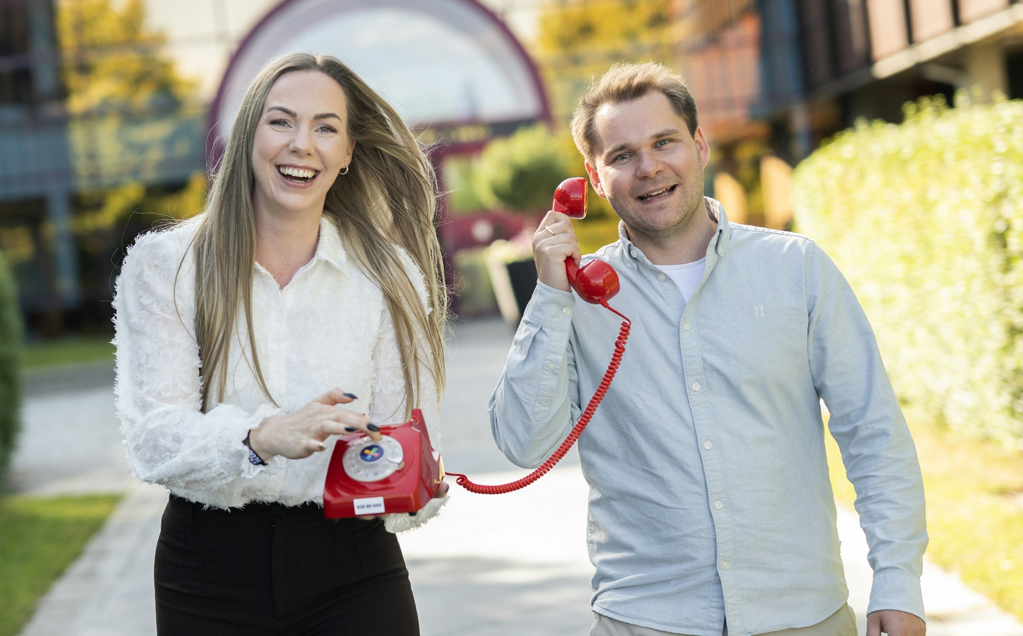 TELEFONEN FRA HAMAR: Magnus Solberg Nygård og Ida Zachariassen i Norsk Tipping er en del av teamet som ringer millionvinnere i Supertrekning.  Bilde av de to utenfor Norsk Tippings inngang. Har med rød telefon. Sommer. Smilende.