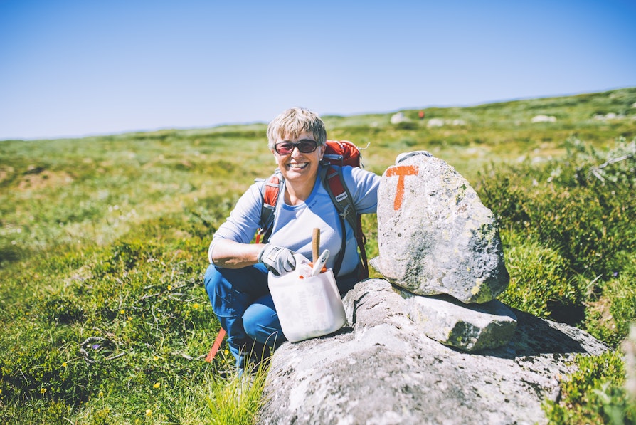 Dame sitter på fjellet ved en stein med en rød T hun nettopp har maltt for å merke sti for DNT.