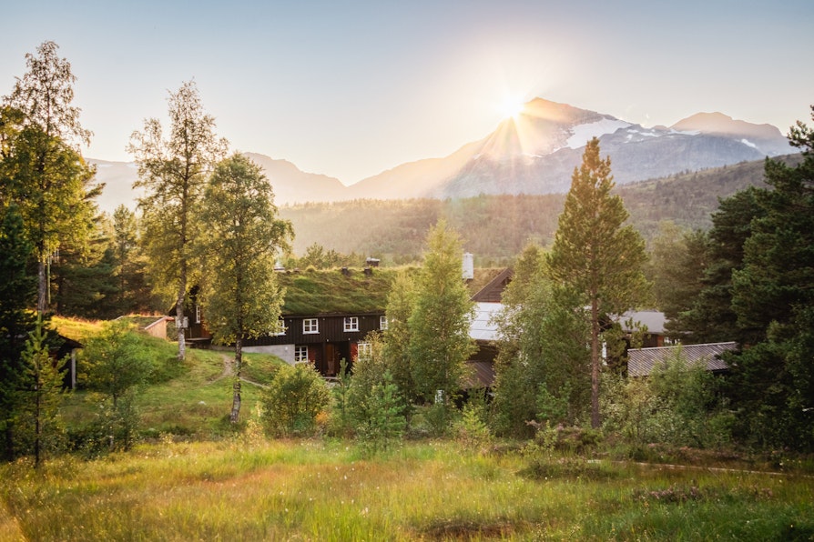 Solen går ned bak et spisst fjell i Trollheimen. man ser en DNT hytte mellom trærne i forgrunnen.