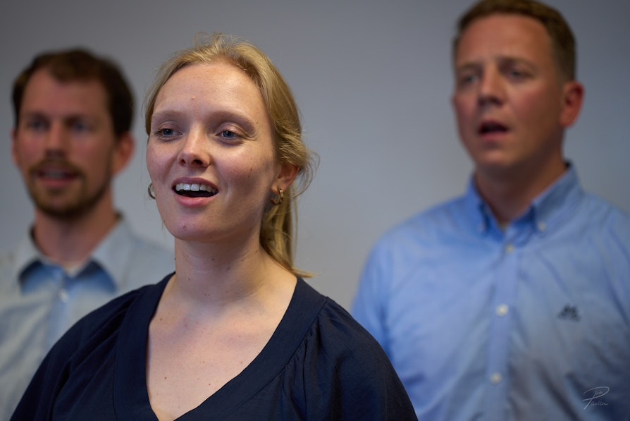 En ung dame synger i et kor. Det er to menn som står bak henne og synger.