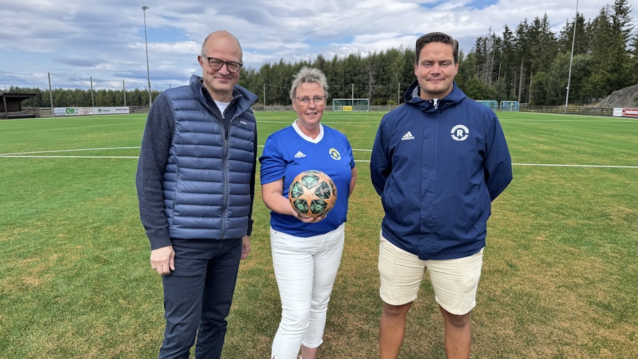 De tre står sidenav hverandre på den nye fotballbana. Inger holder en fotball.