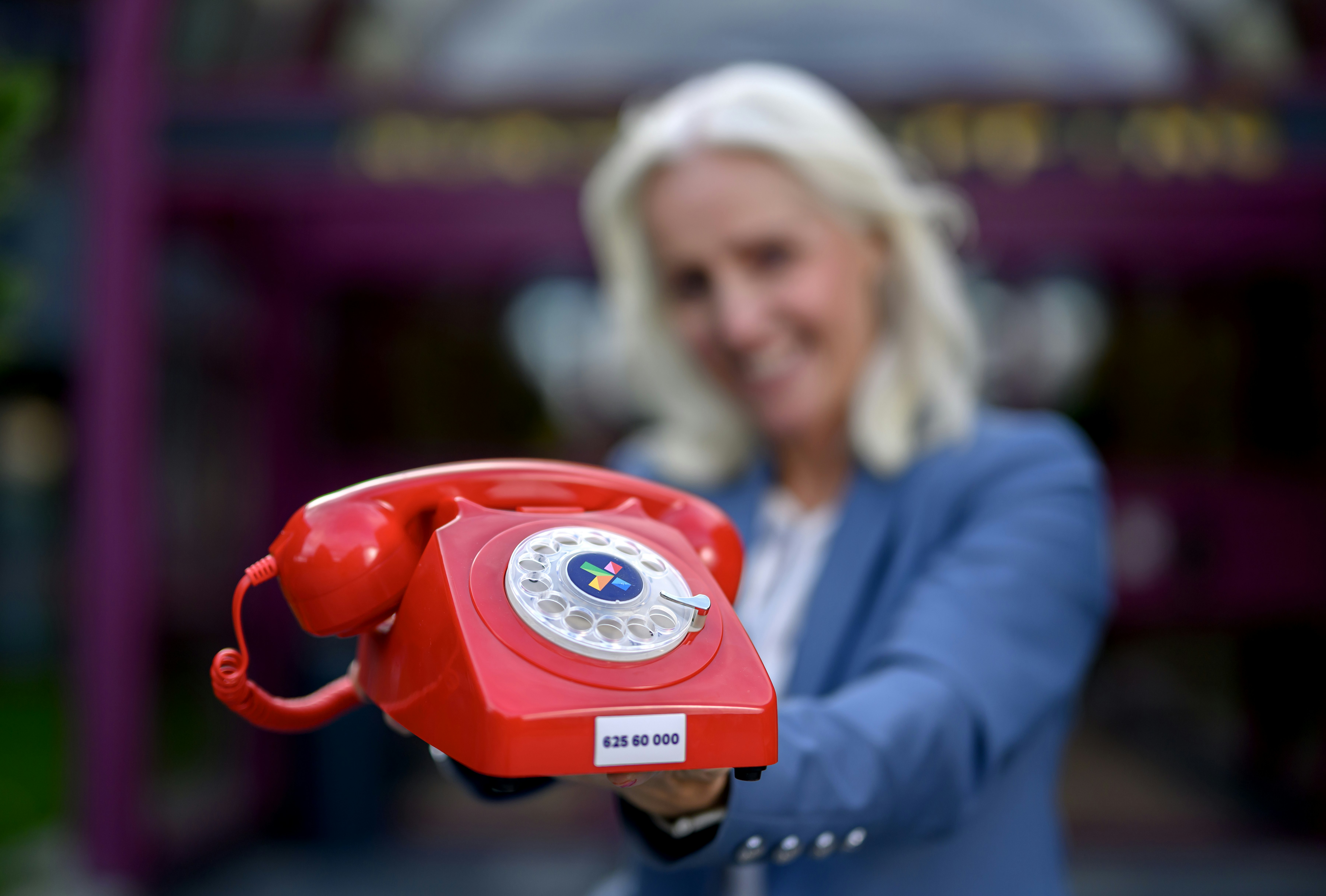 Bilde av kvinne, uskarp, som holder en rød telefon - type gammeldags med nummerskive - foran seg. Bilde tatt i forbindelse med Supertrekning i Lotto høst 2025. Bildet tatt utenfor inngangen til Norsk Tipping, Hamar.