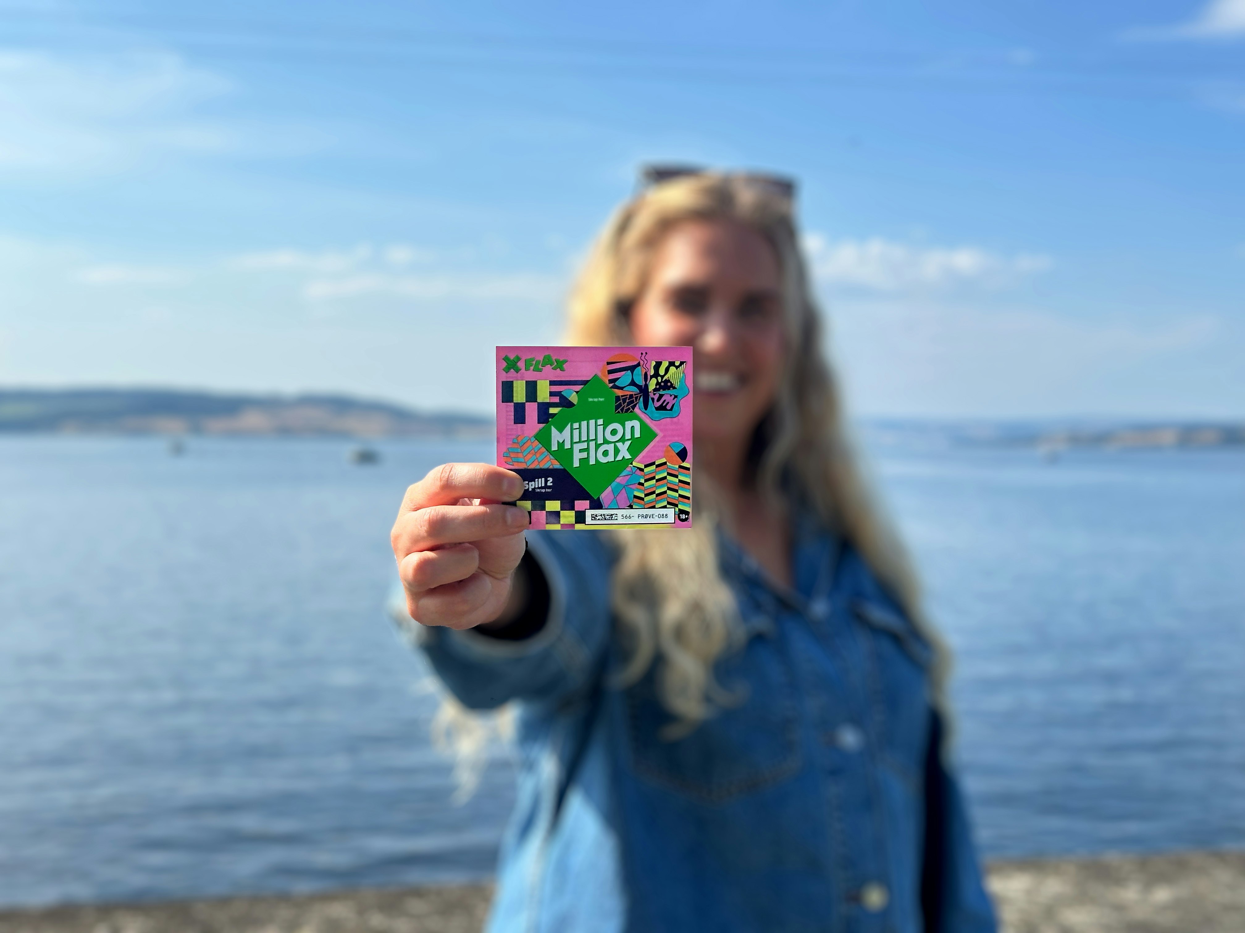 Kvinne med langt blondt hår, solbriller på hodet og ola-skjorte, smiler og holder opp et MillionFlax. I bakgrunnen skimtes Mjøsa om sommeren.