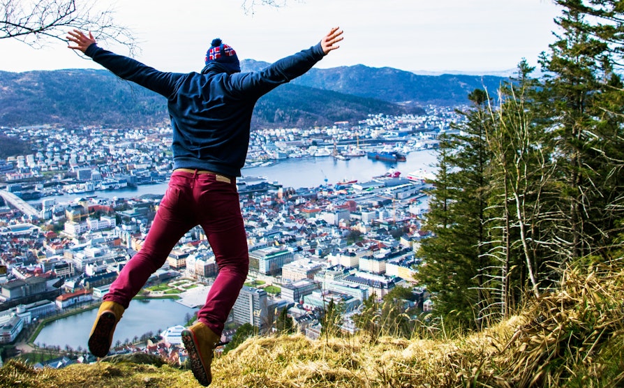 Mann med lue, røde jeans og blå genser hopper på en topp med utsikt over Bergen.