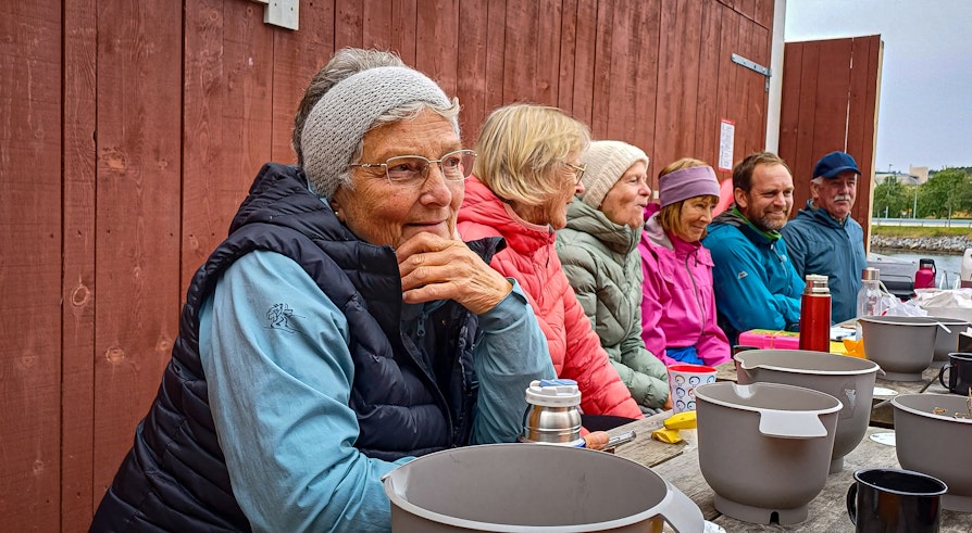 Fem eldre frivillige sitter på en benk på et picnic bord og spiser og drikker og sosialierer. De har på seg godt med turklær.