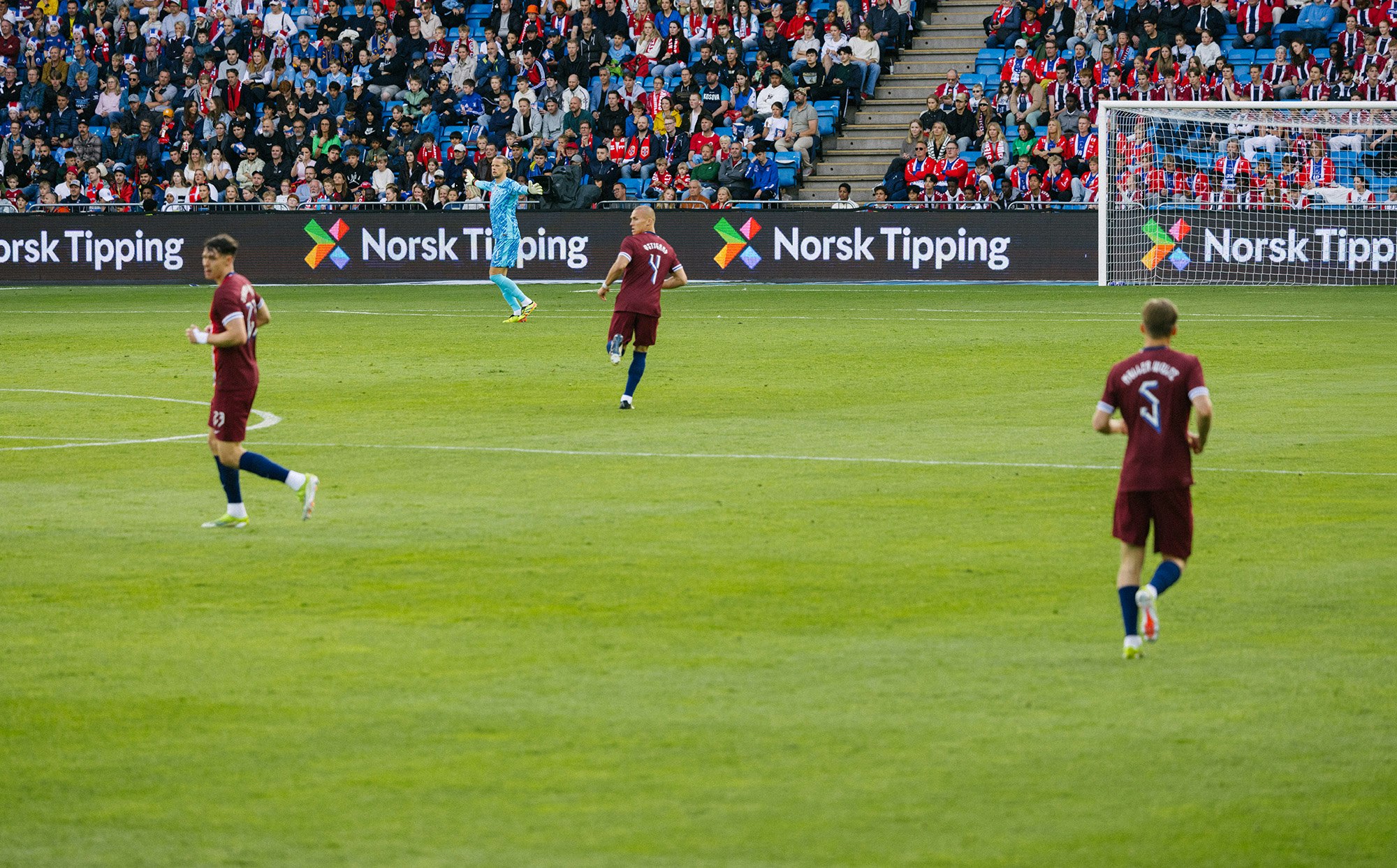 Tre fotball-spillere løper på en fotballbane. Norsk Tipping-logoen i bakgrunnen.