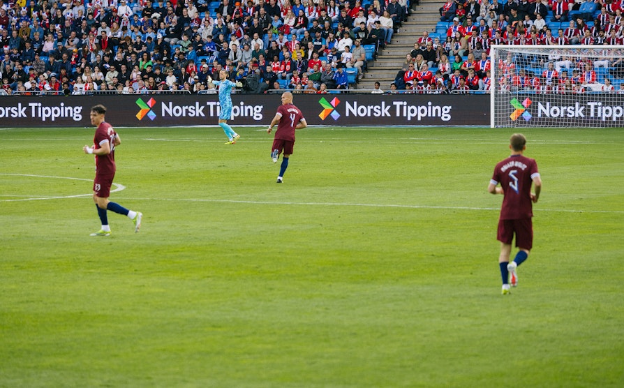 Tre fotball-spillere løper på en fotballbane. Norsk Tipping-logoen i bakgrunnen.
