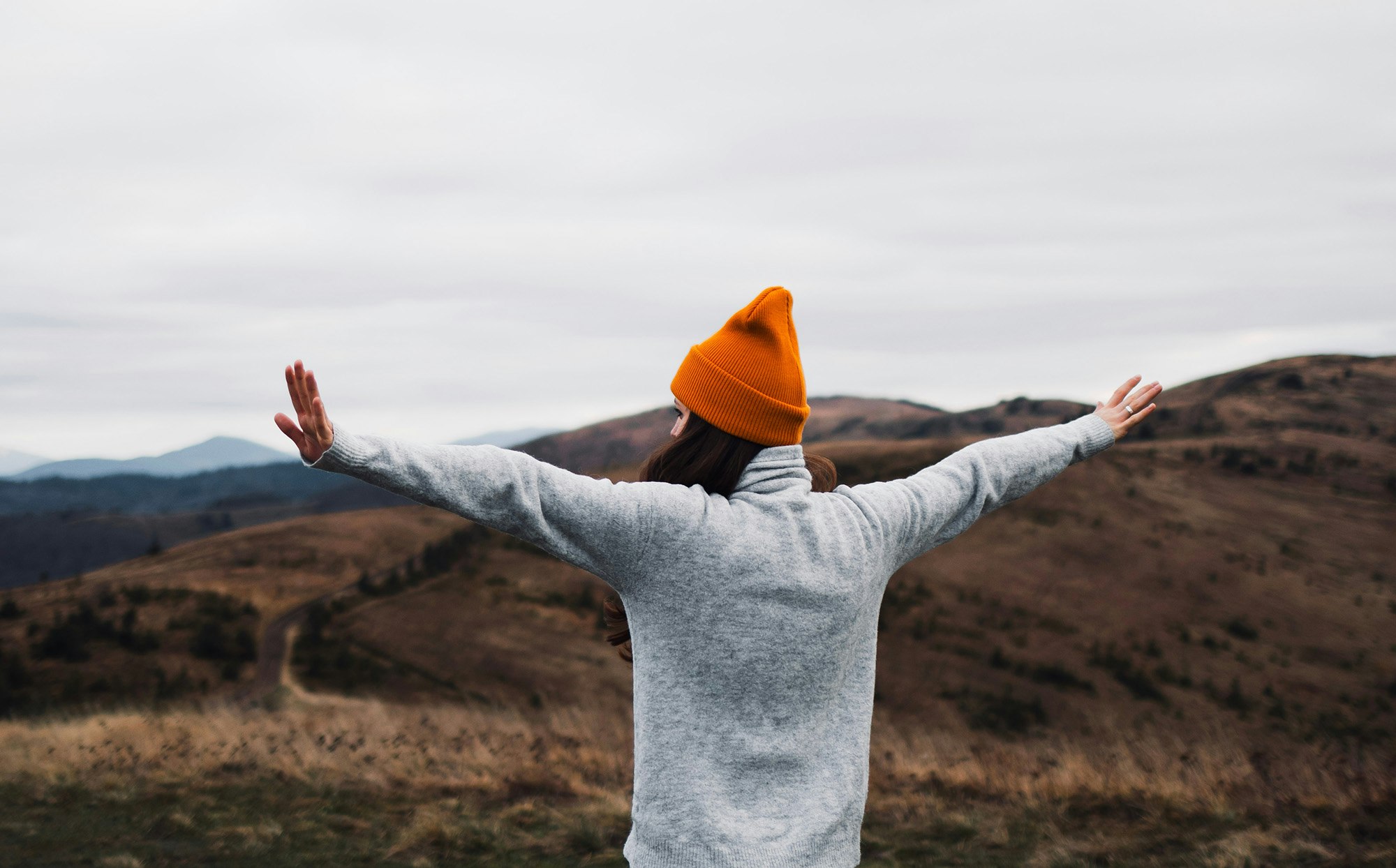 Kvinne med oransj lue bakfra jubler i naturen