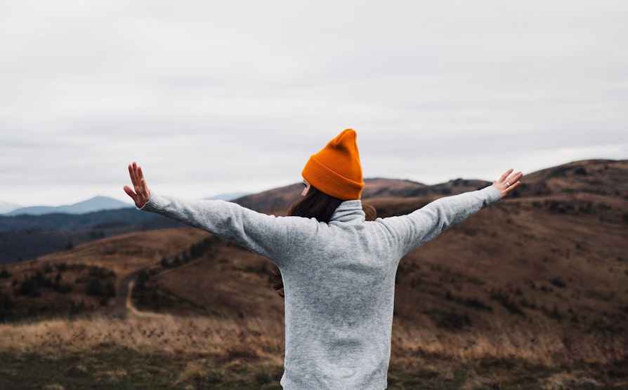 Kvinne med oransj lue bakfra jubler i naturen