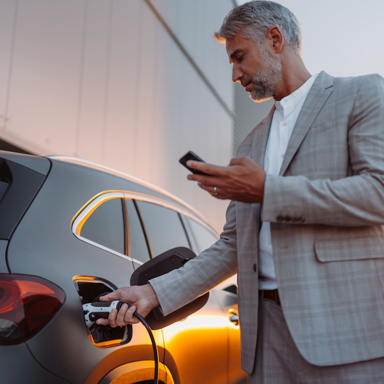 Zakenman houdt smartphone vast terwijl hij auto oplaadt bij oplaadstation voor elektrische voertuigen