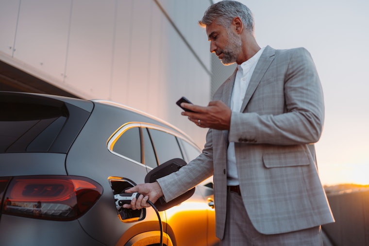 Zakenman houdt smartphone vast terwijl hij auto oplaadt bij oplaadstation voor elektrische voertuigen