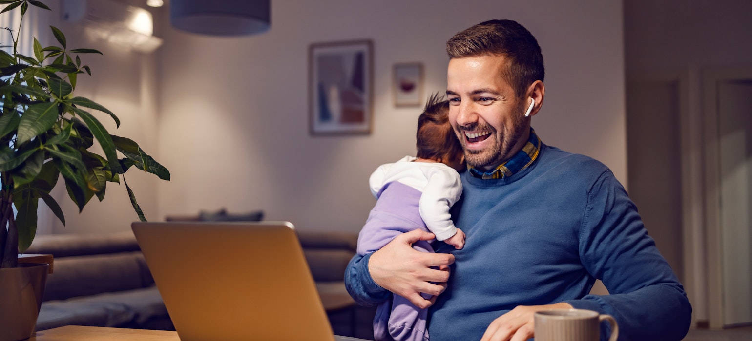 Een gelukkige jonge vader houdt zijn kleine meid in zijn handen en heeft een online vergadering op een laptop met collega's.