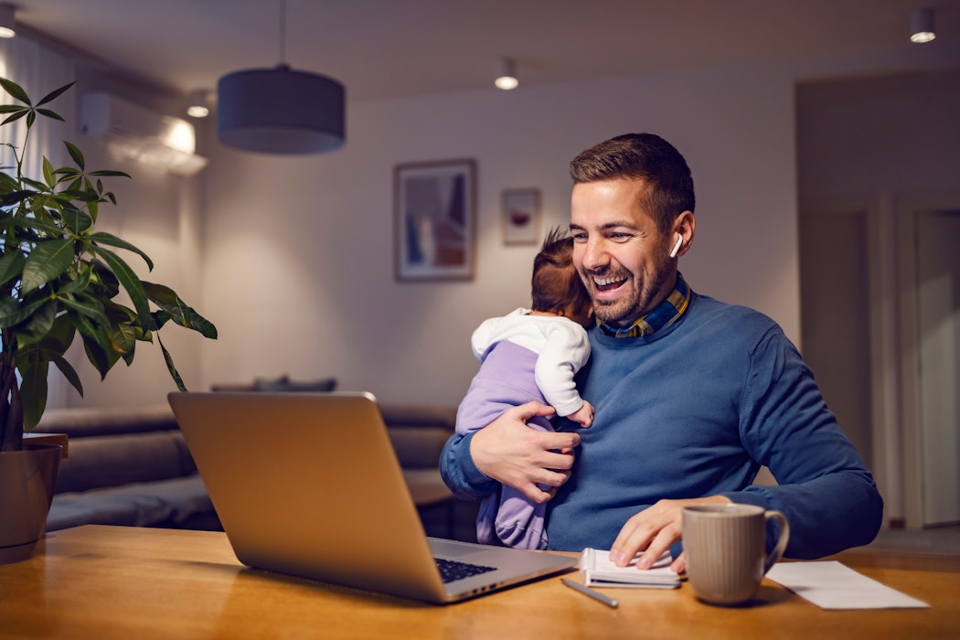Een gelukkige jonge vader houdt zijn kleine meid in zijn handen en heeft een online vergadering op een laptop met collega's.