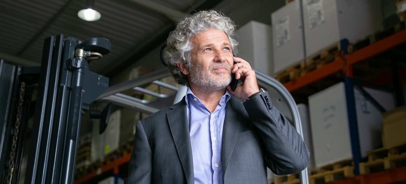 volwassen zakenman die bij een vorkheftruck in een magazijn staat en met een mobiele telefoon spreekt.