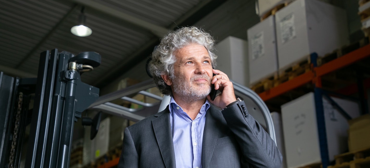 Homme d'affaires adulte debout près d'un chariot élévateur dans un entrepôt, parlant sur un téléphone portable.