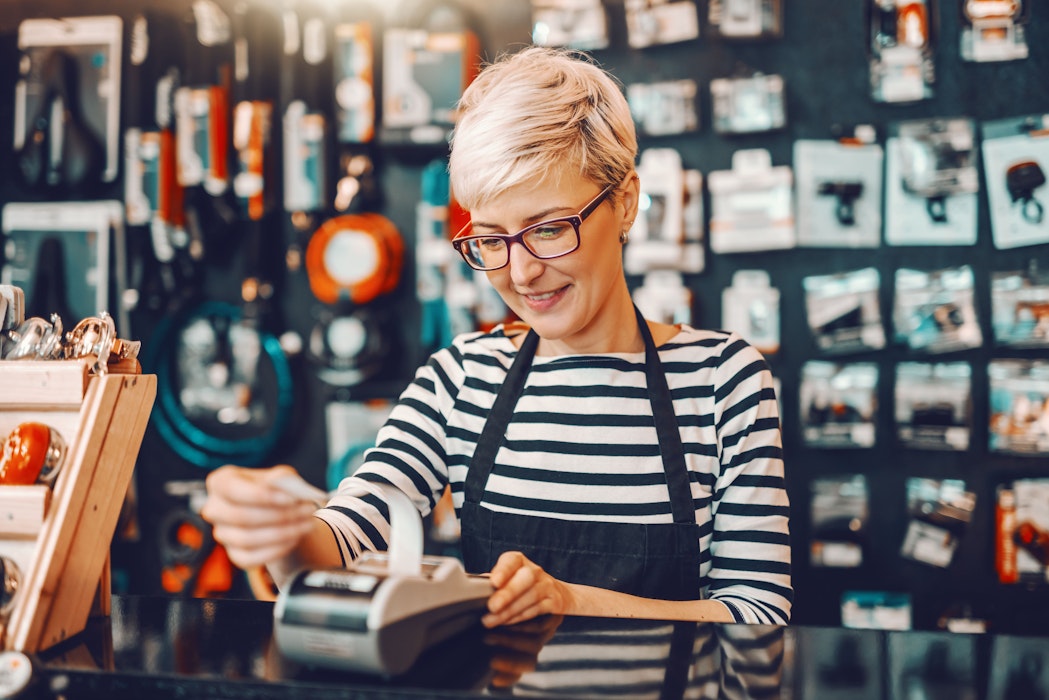 vrouwelijke werknemer met kort blond haar en een bril gebruikt de kassa terwijl ze in een fietsenwinkel staat.