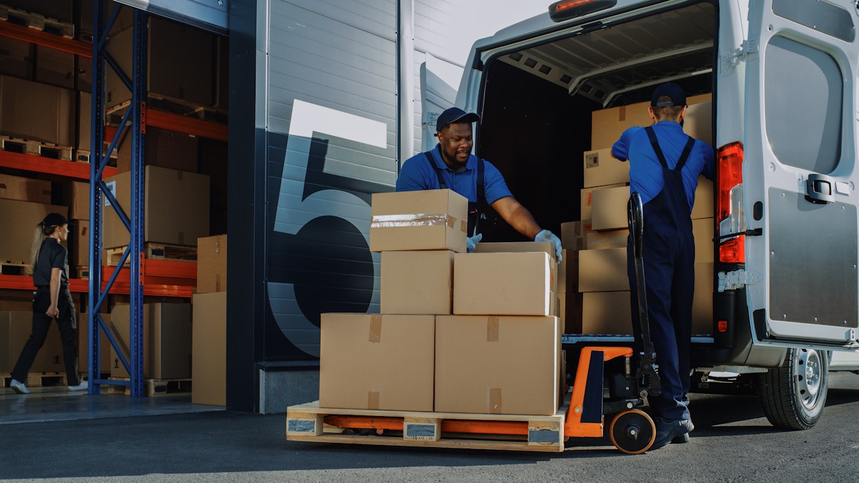 Buiten aan een logistiek magazijn met een open deur wordt de bestelwagen geladen met kartonnen dozen.