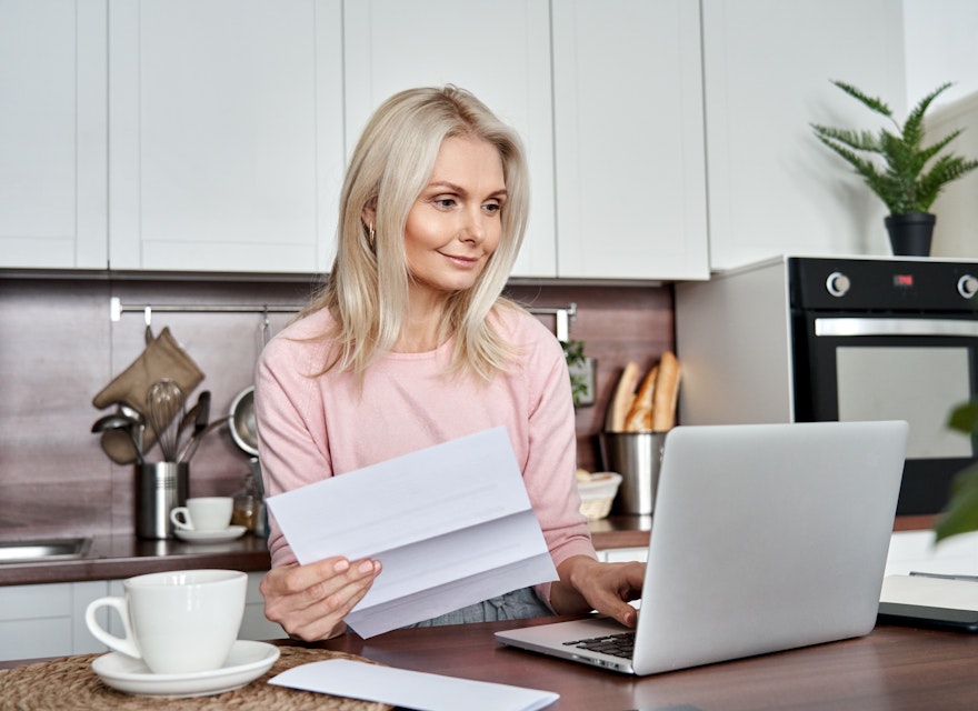 Vrouw kijkt naar laptop met papieren in de hand