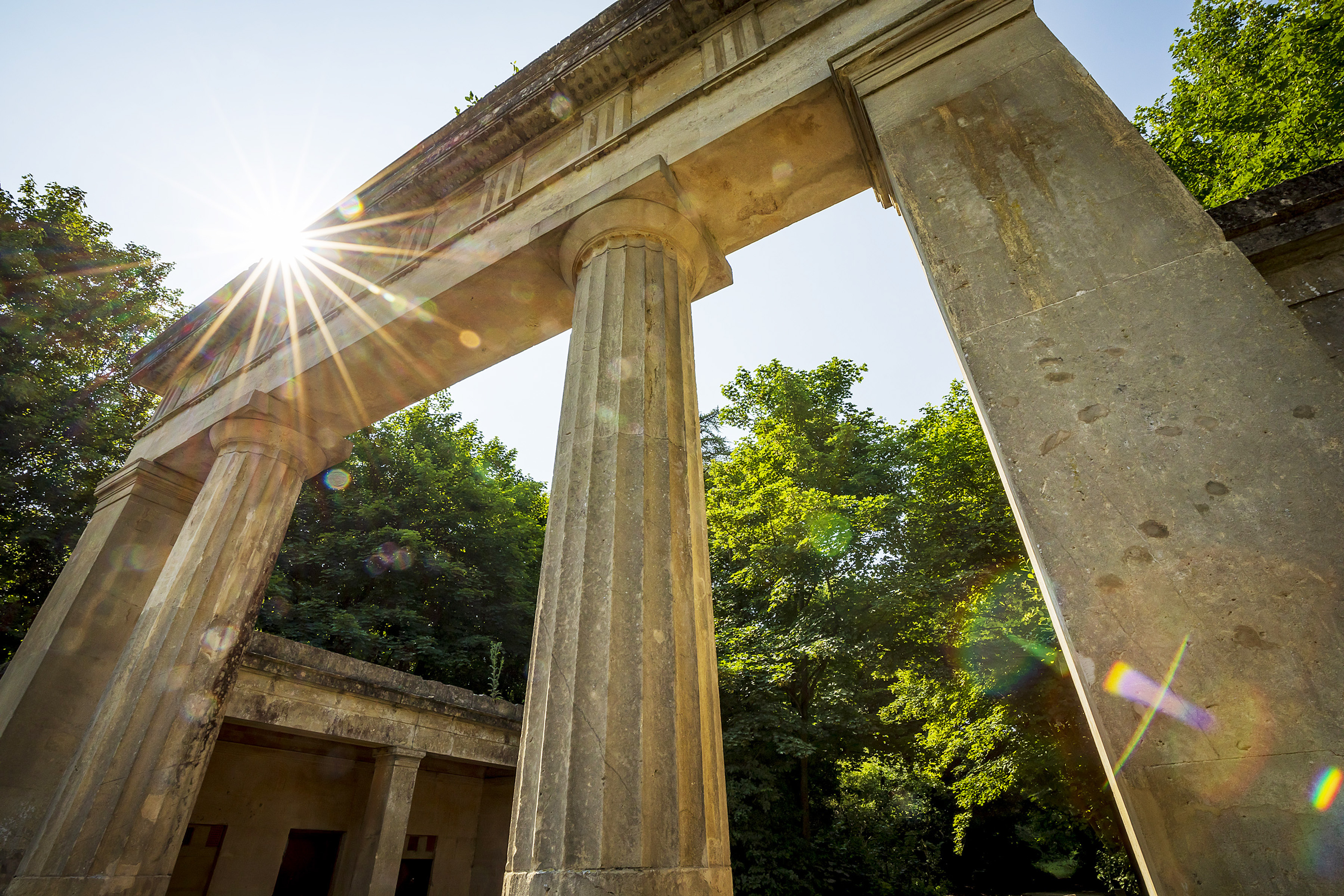 The picture is taken looking up at the stone columns and mantel piece of the entrance to crabtree plantation. In the background you can see green trees. The sun reflect off the top of the structure creating a glare.