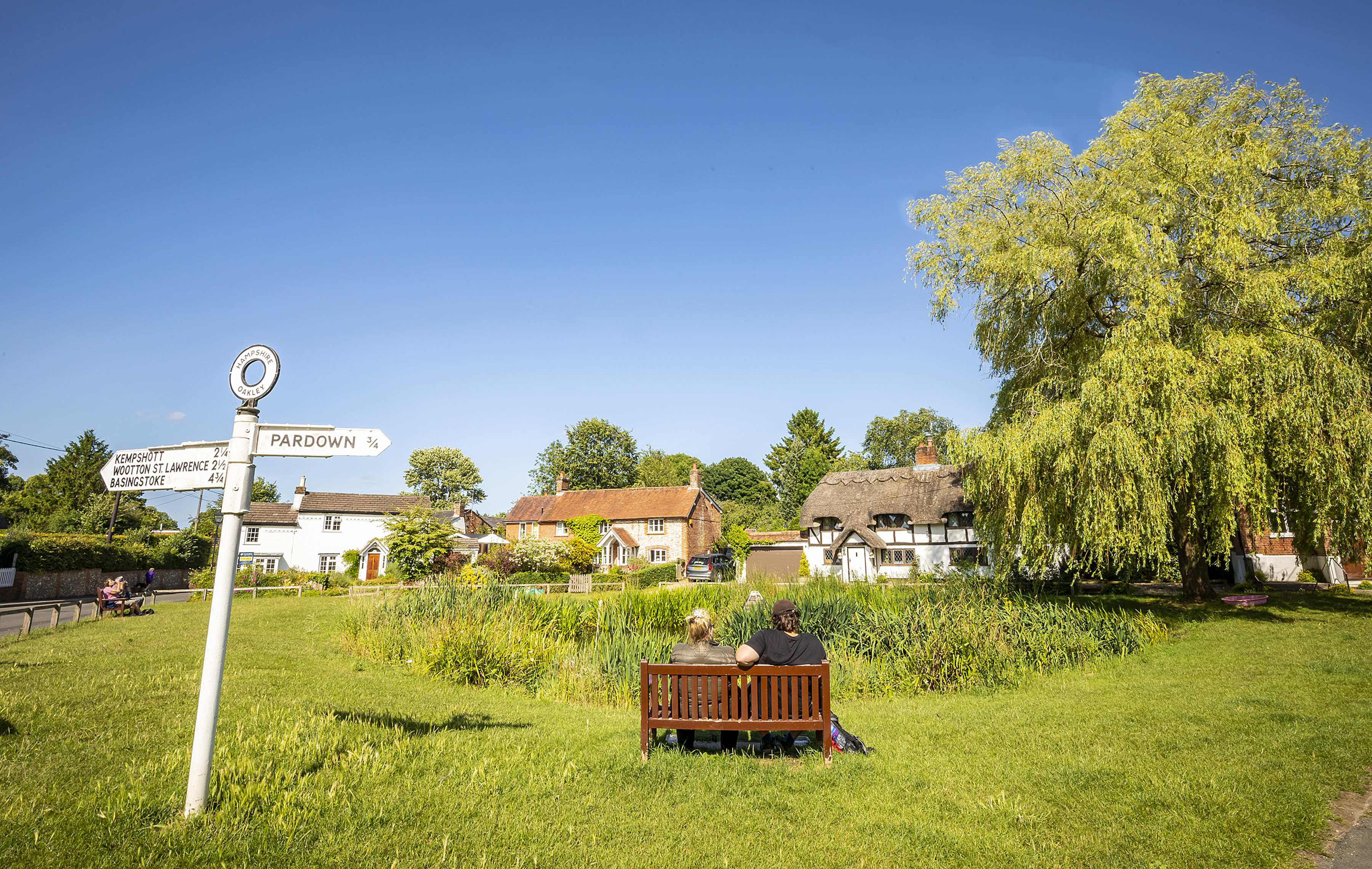 Two people sit on a bench looking out at a pond and the quaint houses of Oakley Village.