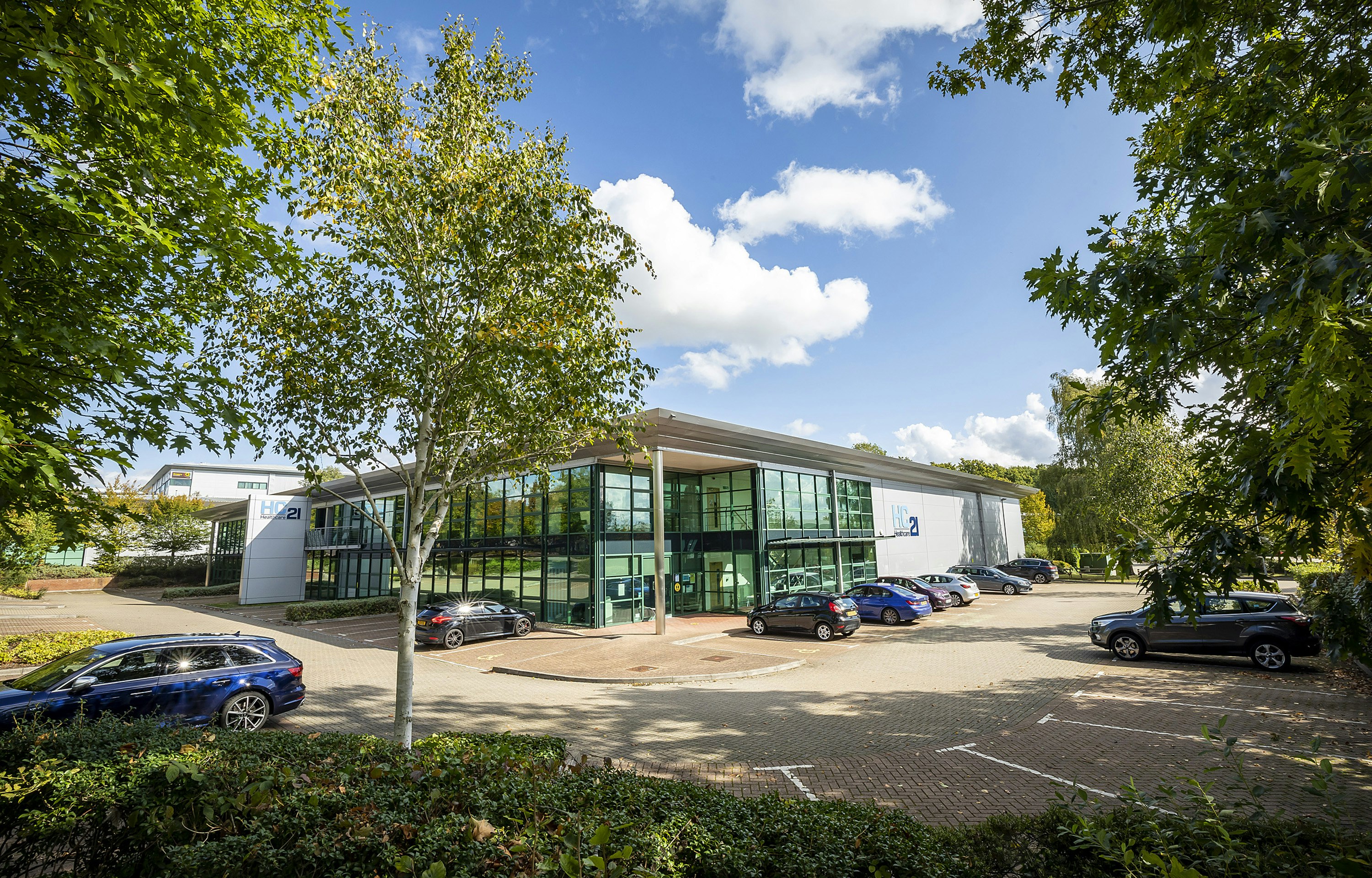 A glass and panelled two storey office building with a car park and trees in front