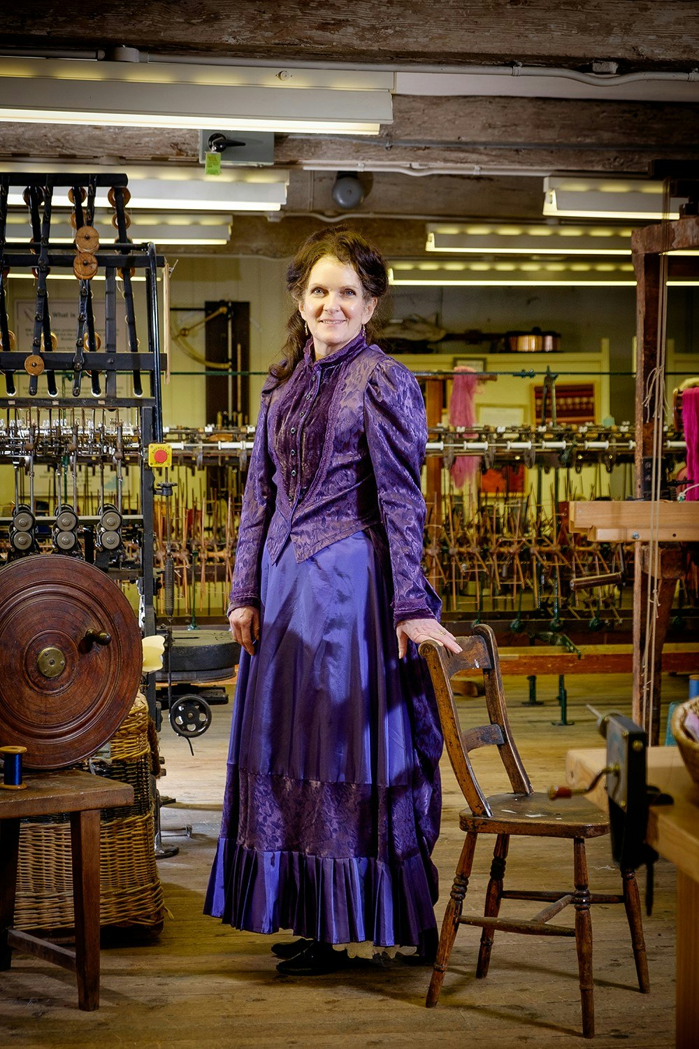 A lady dresses in a purple victorian outfit standing with a hand resting on a chair and with weaving equipment in the background.