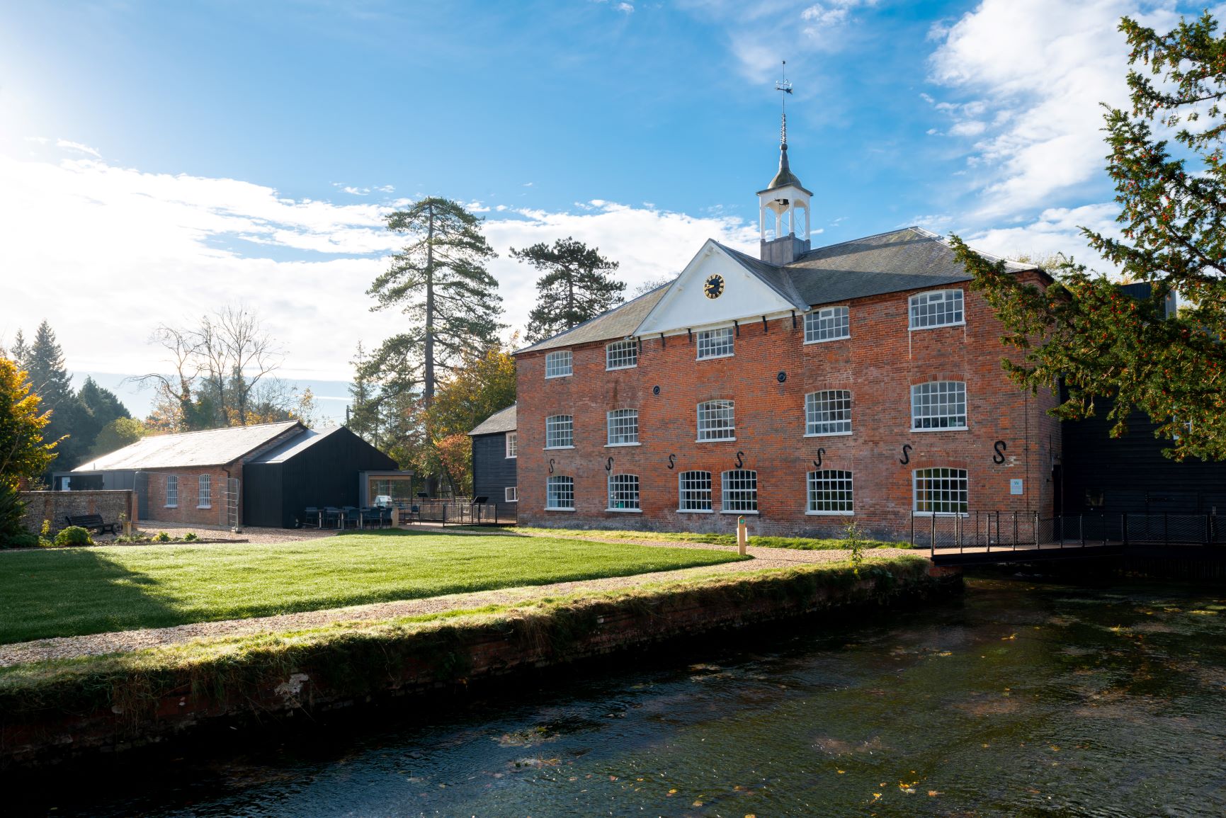 A red three storey brick building with white windows. It has a triangular pitch in the middle of the roof and a tower. To the right of the image is the river.