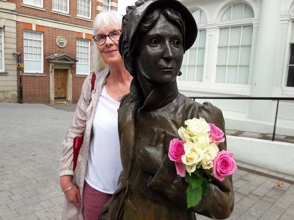 A lady stands behind the statue of Jane Austen who is holding a bunch of roses.
