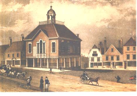 A print. A town hall with columns and a high pitched roof and turret on the top. other houses surround it. There are people and horses and carts on a road in front of them.