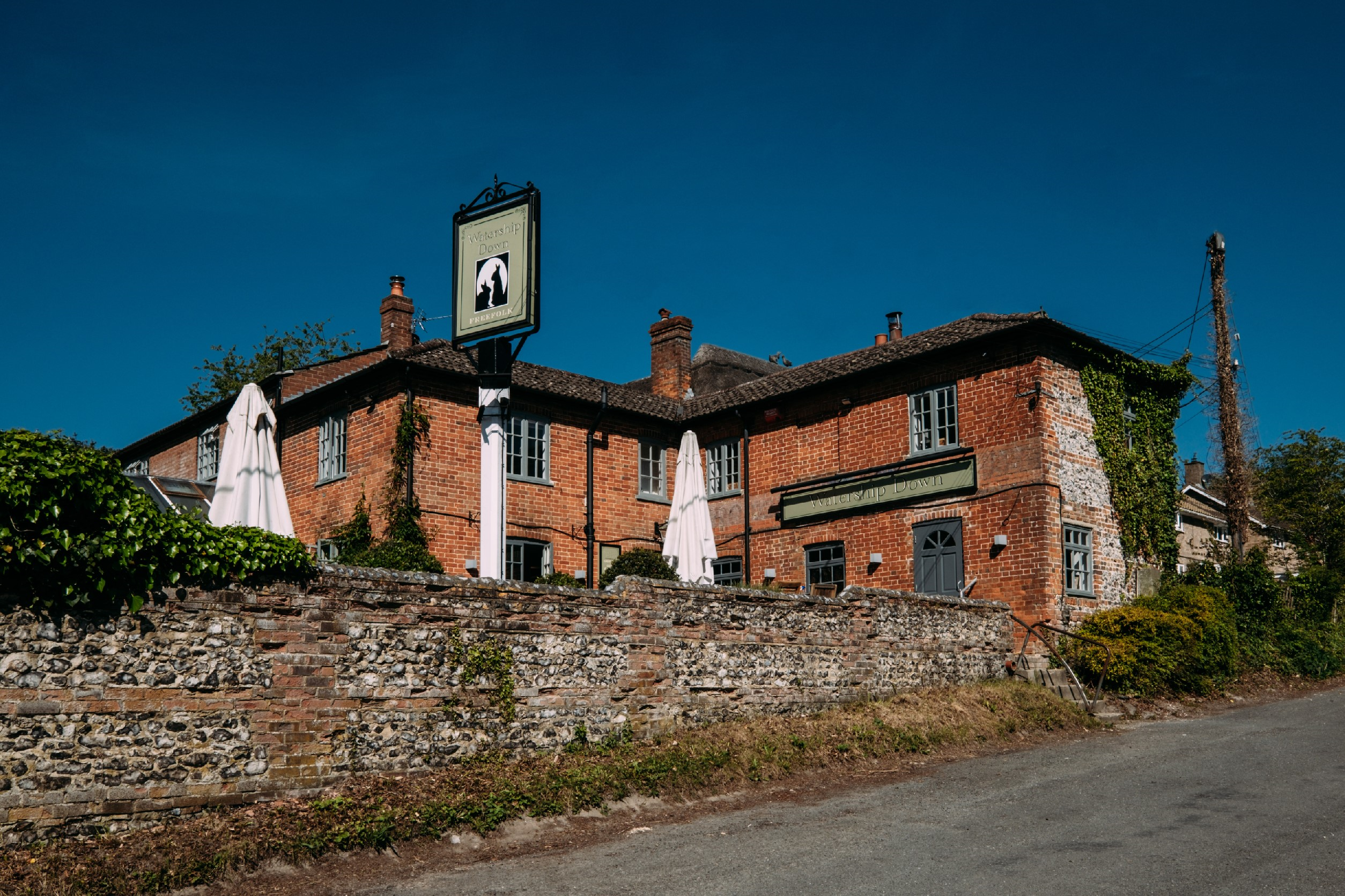 The outside of Watership Down Inn, a red brick two storey building with a stone wall in front. Popping out above the wall you can see two umbrellas which are down and the pub sign.