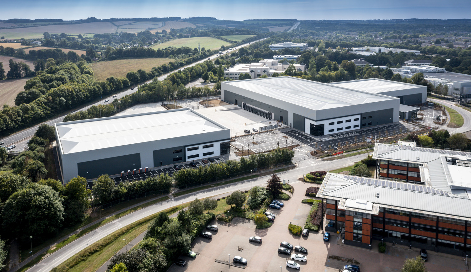 An industrial park from the air. Three large buildings and car parks. In the distance you can see the motorway and green fields.