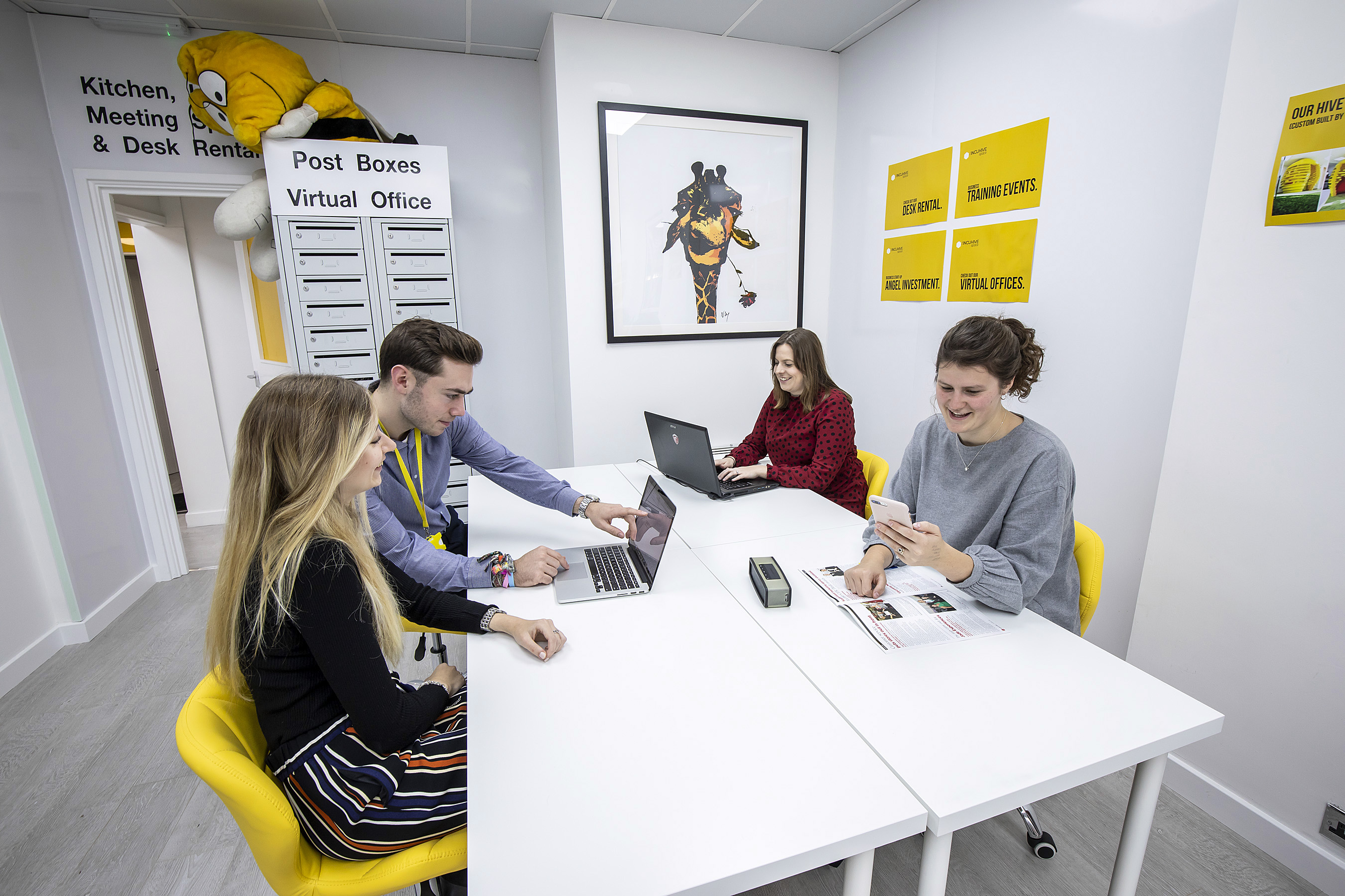 a group of business people sit around a table. One is on a laptop, one is looking at her phone and two are working together looking at a laptop