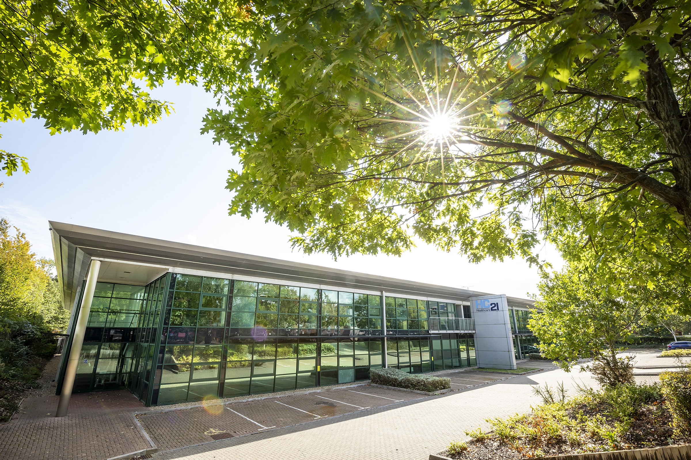 A glass two storey building with overhanging roof. Outside are parking spaces and trees overhang the image