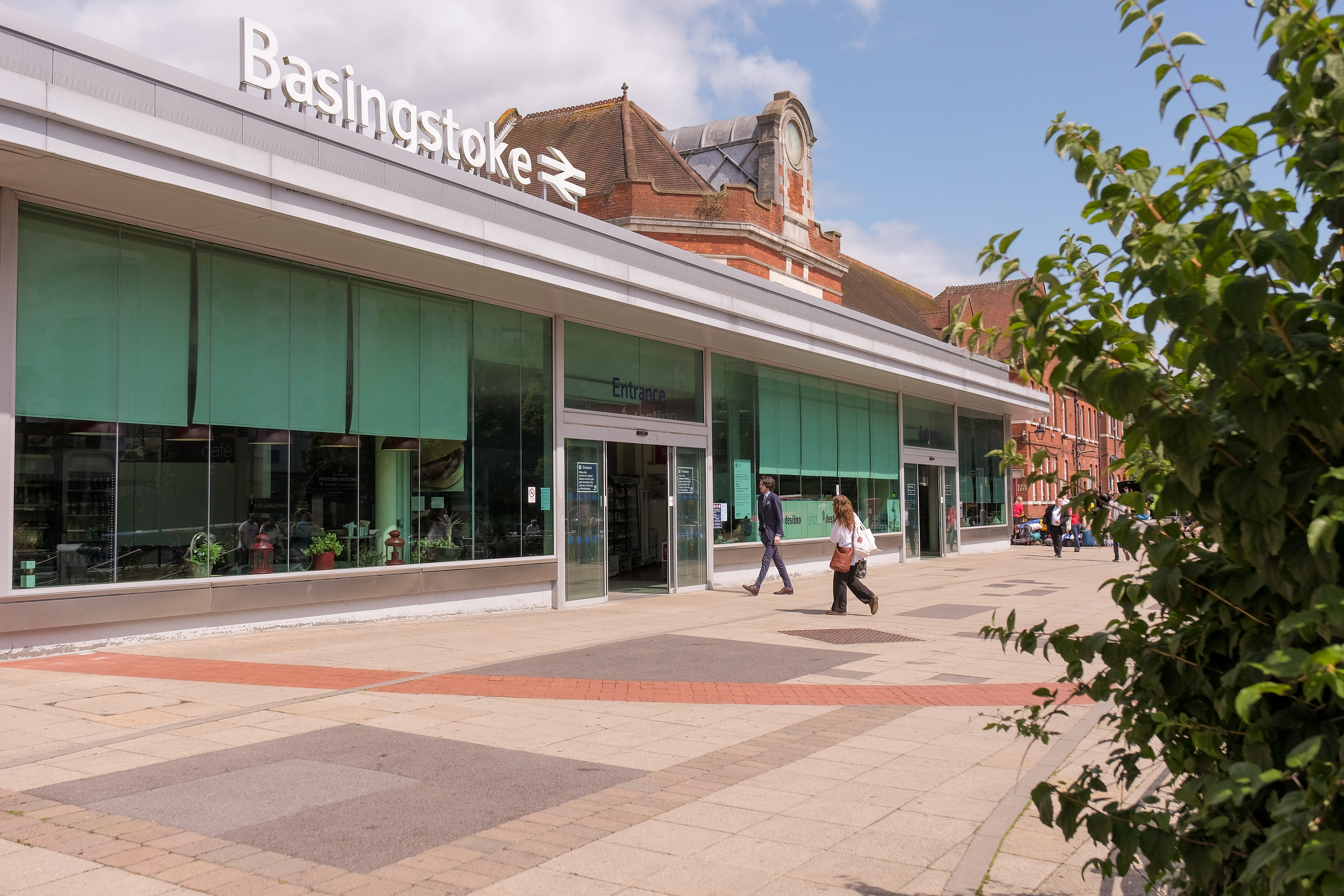 The glass panelled frontage of the train station. A few people are walking in. On the top of the building is the text Basingstoke with the railway sign.