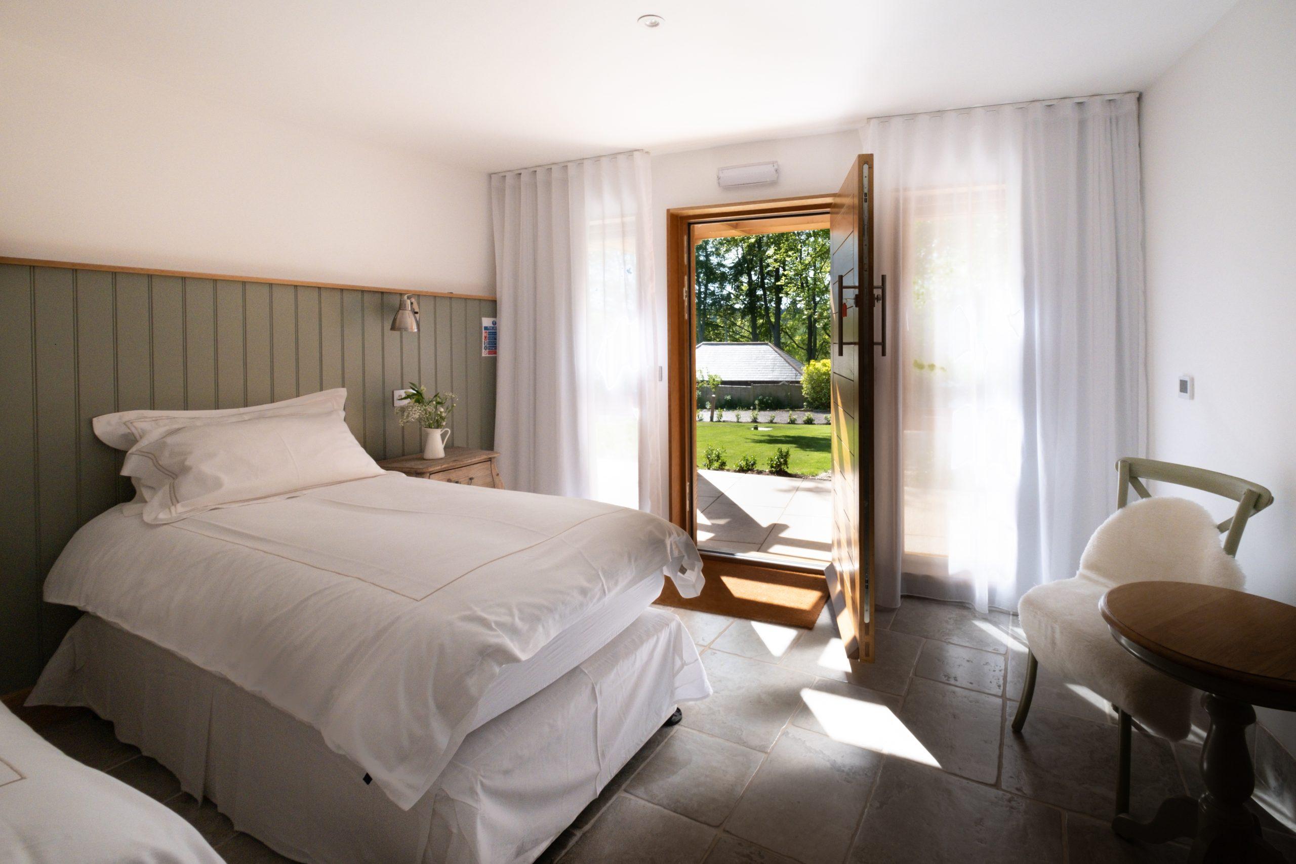 A hotel room with a door to outside to the garden. There is a bed with crisp white linen, a bedside table and a small table and chair opposite.
