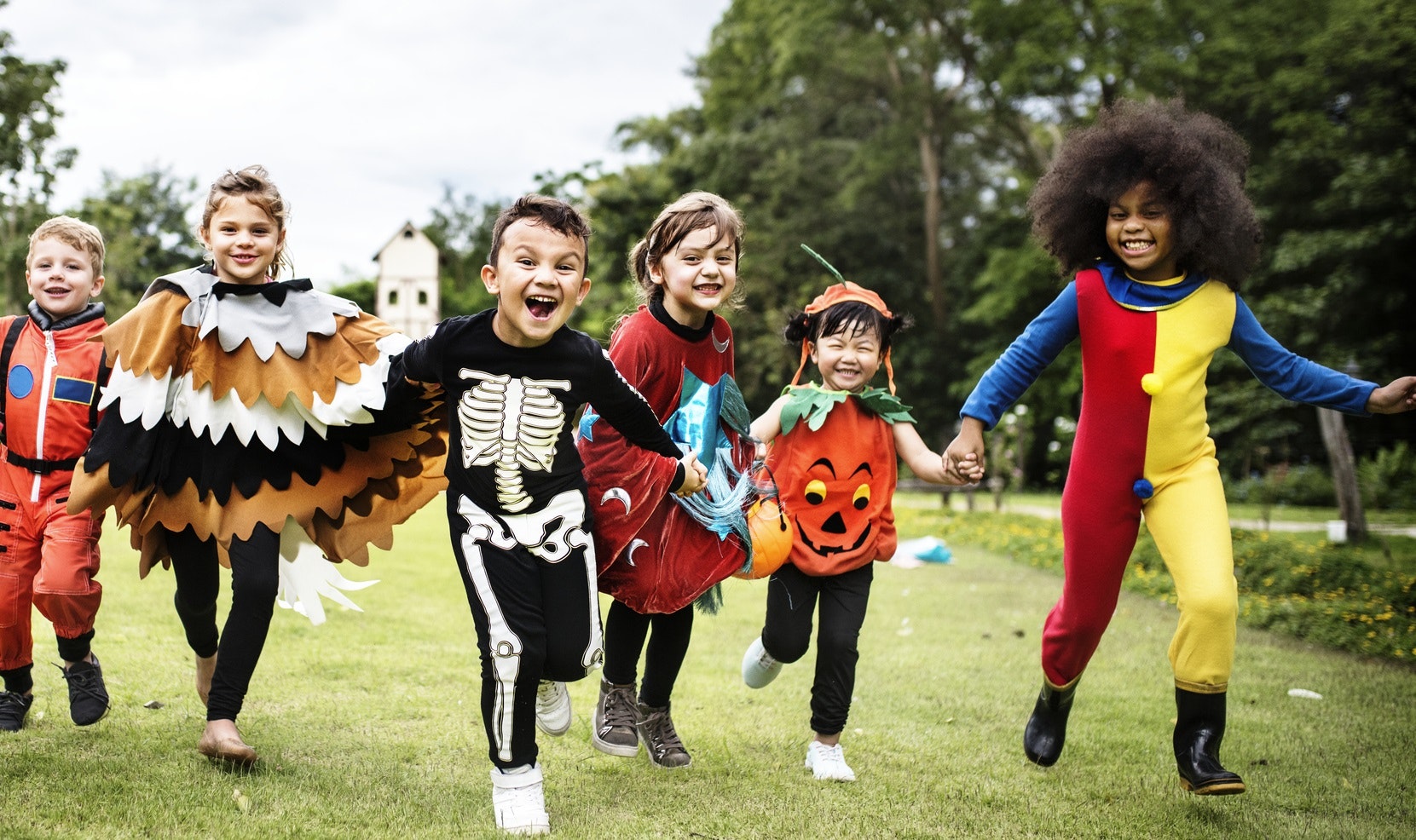 A group of kids running towards the camera wearing Halloween costumes, such as clown, pumpkin, skeleton,. astronaut