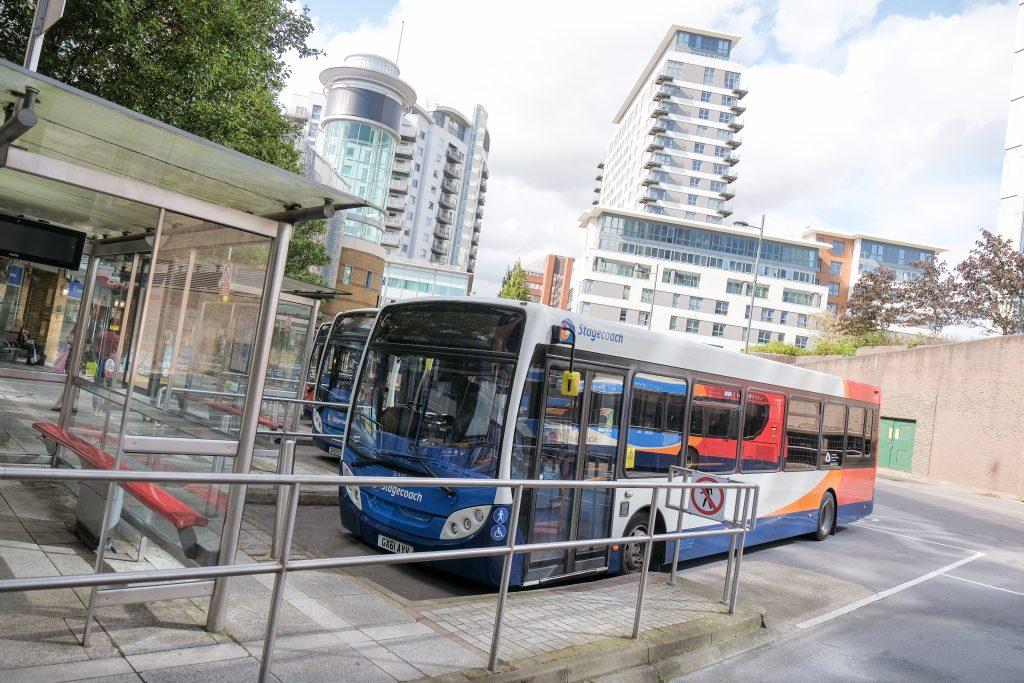 A bus with another behind it is parked up at a bay in the bus station. You can see the skyline of Basingstoke town centre behind it.