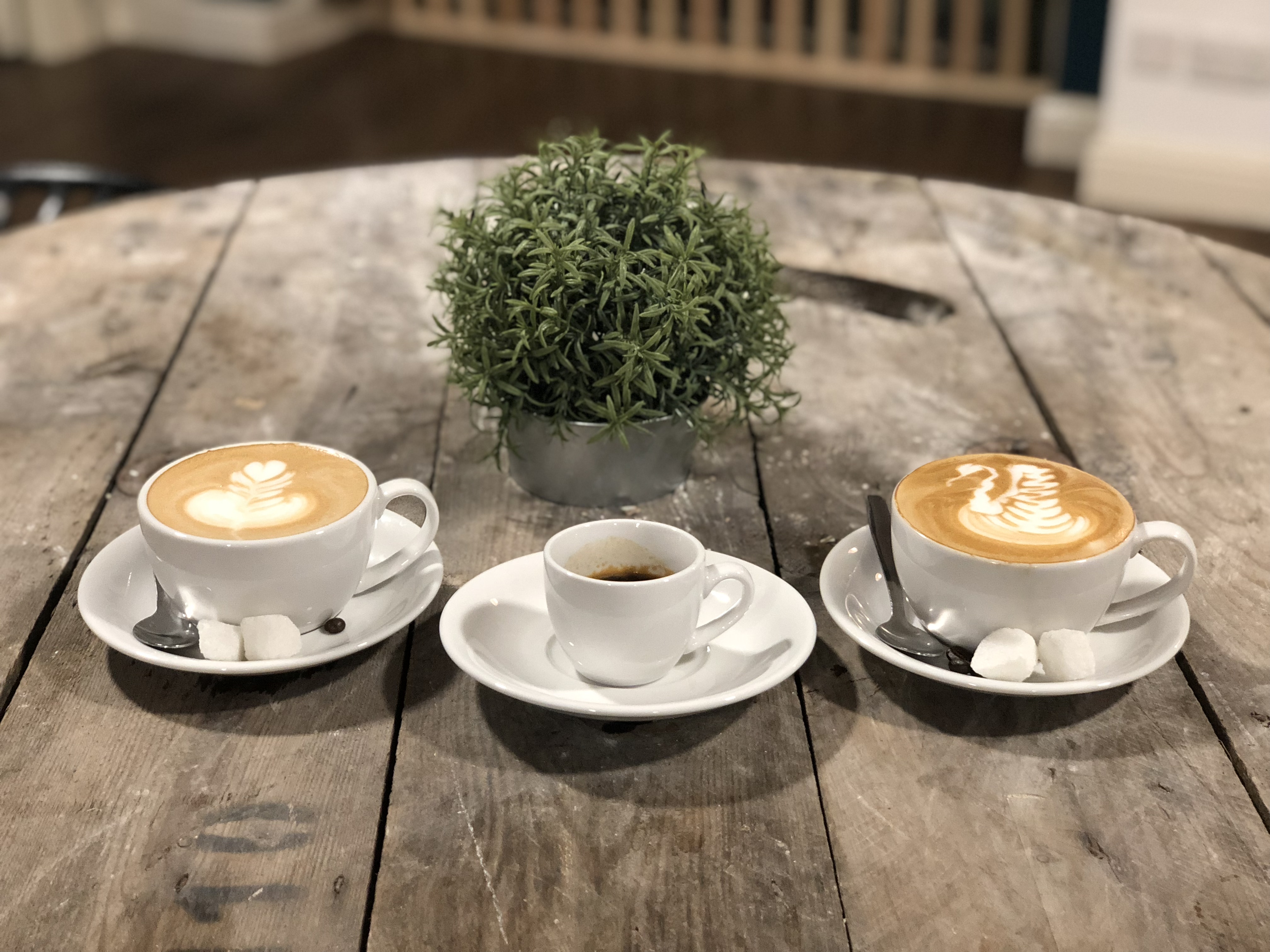 Two regular sized cups of coffee with milky froth on top and a smaller expresso cup in the middle. They sit on a wooden table.