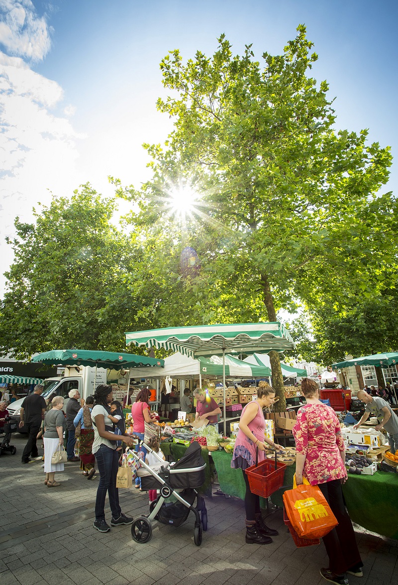 A bustling market with stalls and lots of people. Behind are tall trees.