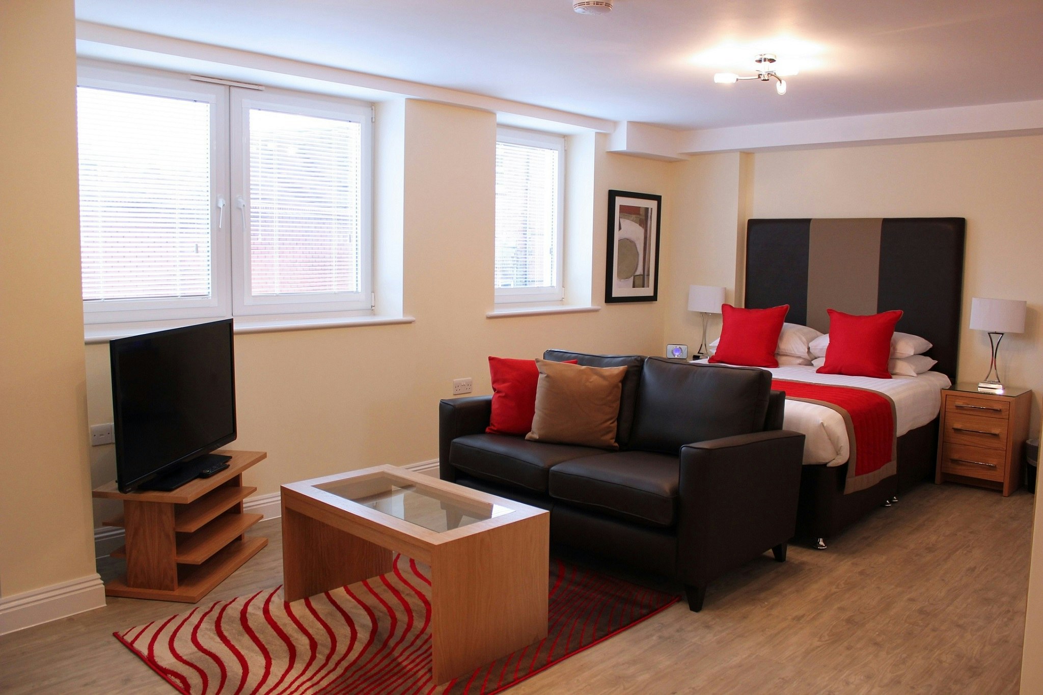 A double bed with white linen and red cushions with two bedside tables. A sofa sits at the end of the bed with a coffee table in front and a tv and tv stand in the corner of the room.