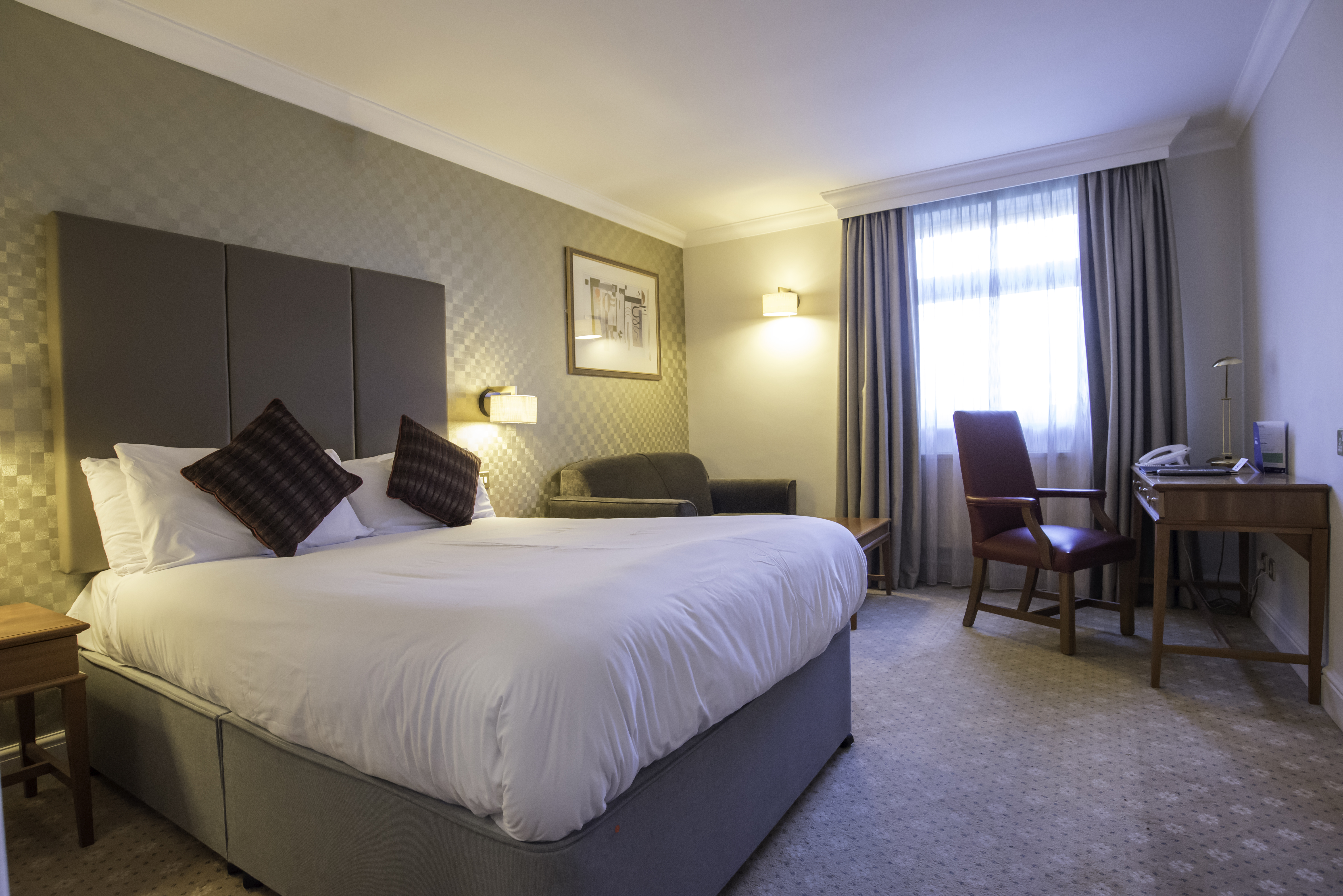 a double bed with crisp white bedding sits in the middle of the room. It has a bedside table one side of the bed a sofa and coffee table the other side of the bed. Opposite is the desk and a chair. At the far end is a window.