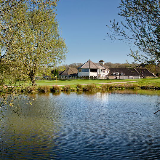 A white clubhouse building sits aside a picturesque lake. The sky is blue.