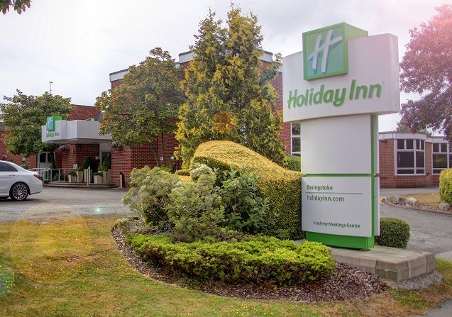 A white and green Holiday Inn sign. It is surrounded by green shrubs. In the background you can see the hotel.