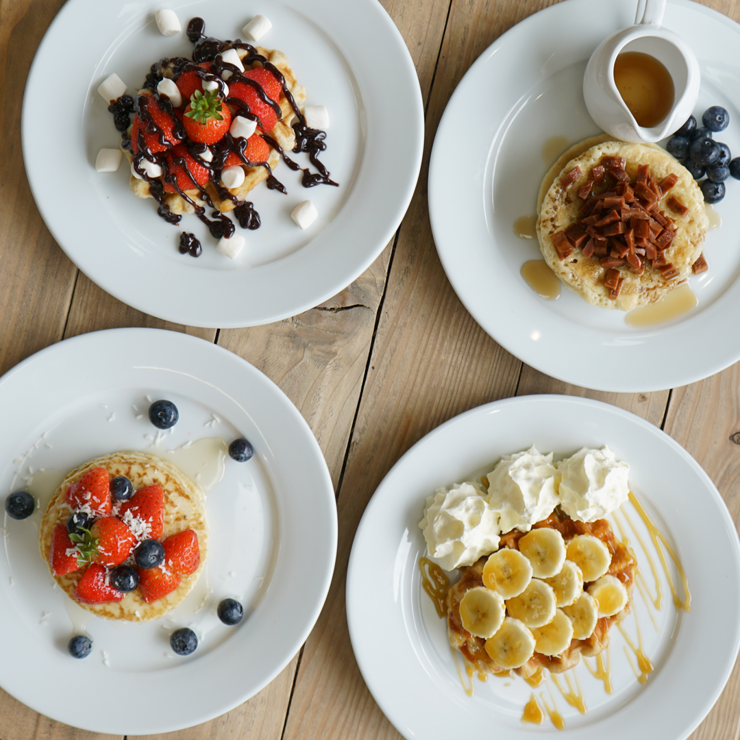 Four white plates full with various pancake and waffle receipes. Including berries, banana and syrups.