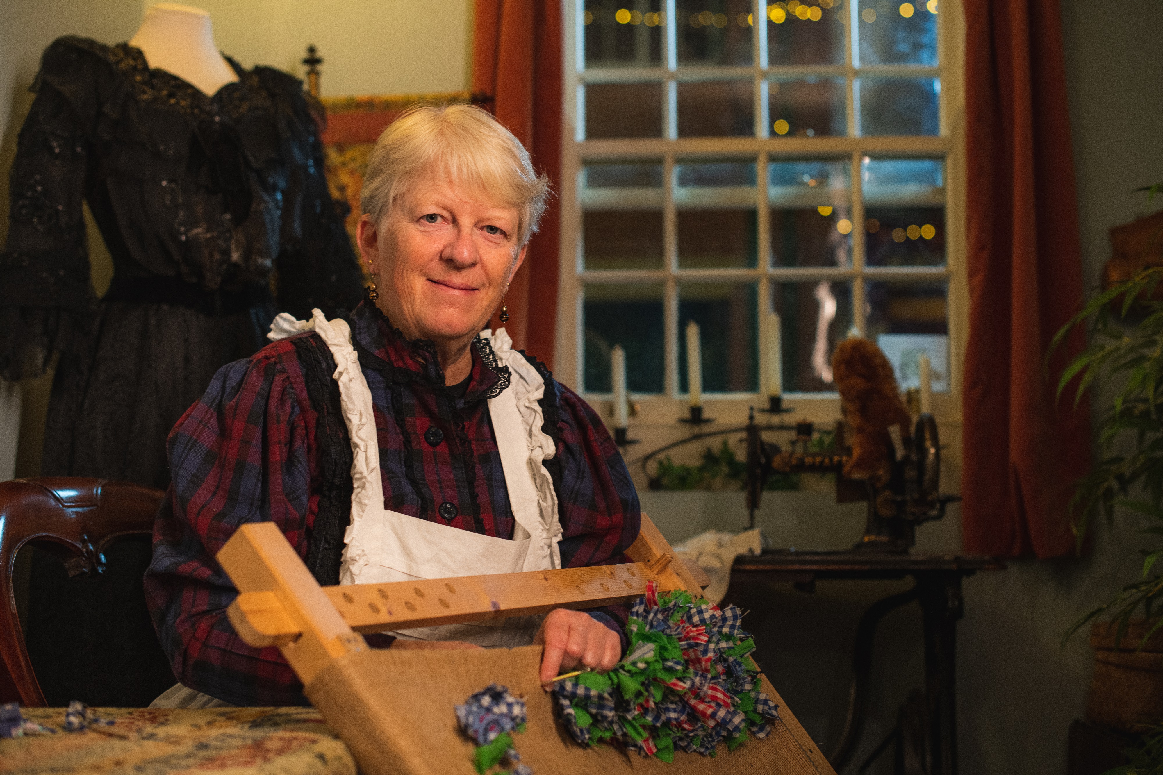 A lady with grey hair smiles at the camera. She is wearing Victorian era clothing, a checked red and green dress with a white apron. She is weaving.