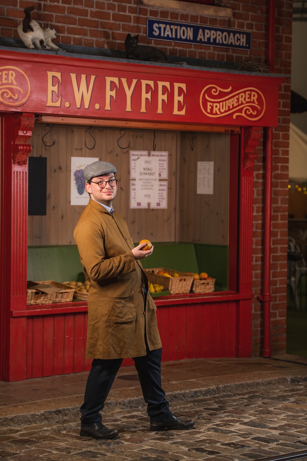 A man dressed in a brown overall and flat cap stands infront of a Victorian shop front holding an apple.